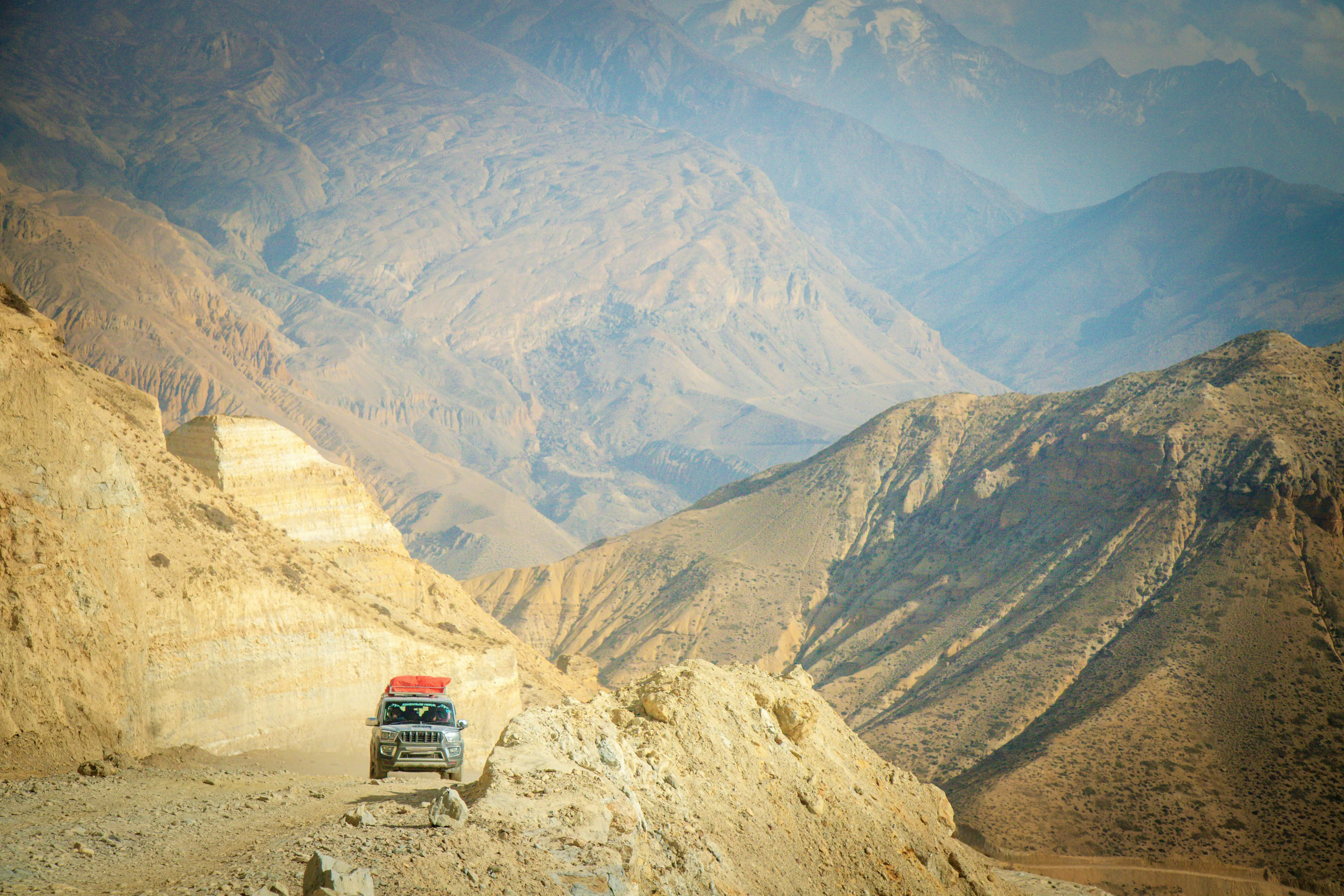A vehicle driving on a mountain road towards Muktinath Temple, with rugged terrain and Himalayan landscapes in the background.